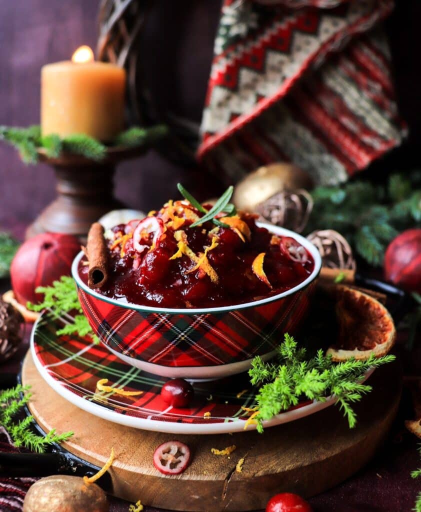 A festive bowl of honey orange cranberry sauce topped with orange zest and rosemary, set beside a lit candle and holiday greenery on a rustic table.