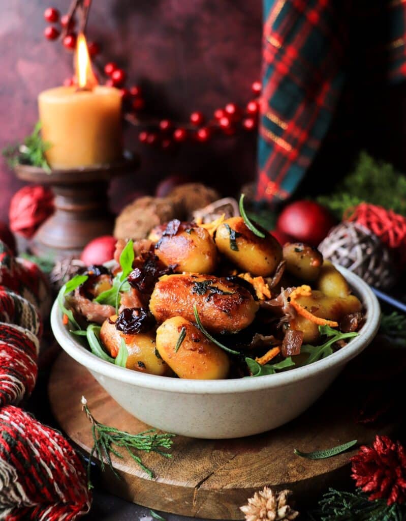 Golden pan-fried gnocchi with cranberries, prosciutto, and citrus zest in a bowl, styled with warm candlelight and Christmas greenery.