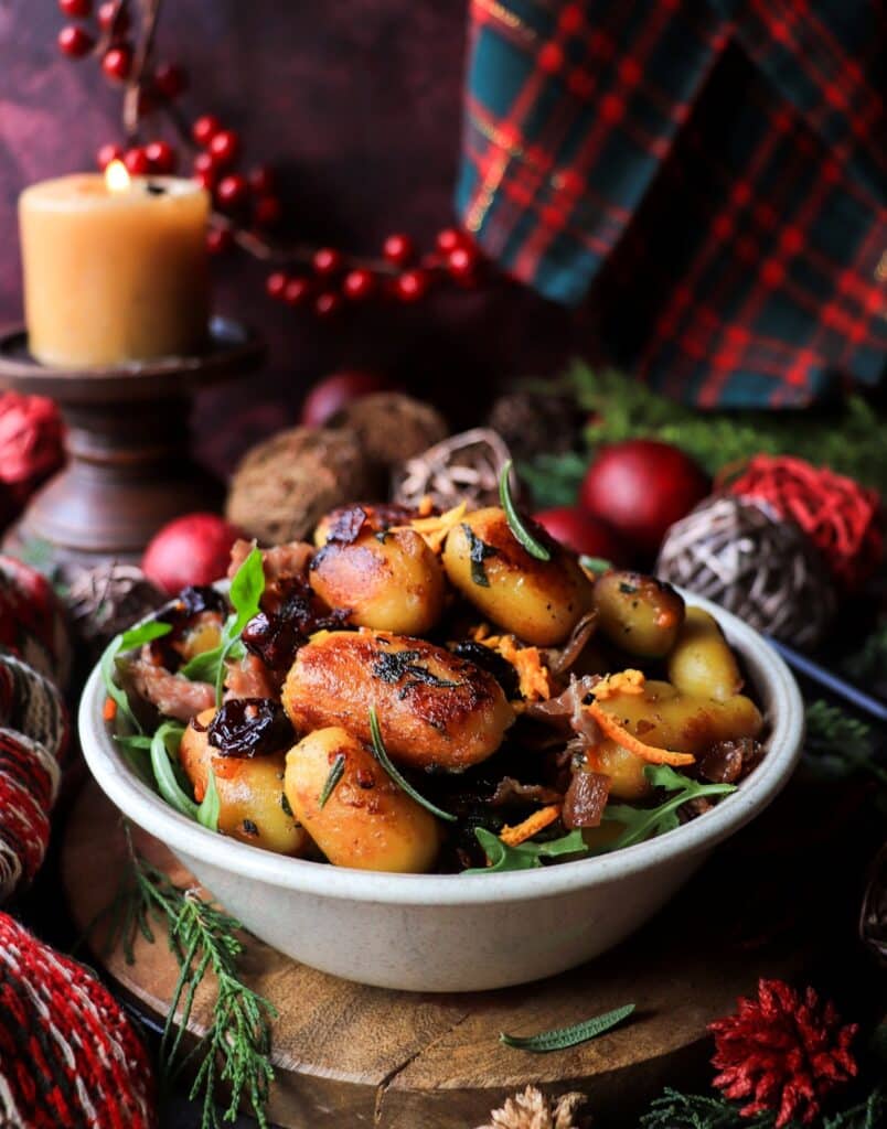 Golden pan-fried gnocchi with cranberries, prosciutto, and citrus zest in a bowl, styled with warm candlelight and Christmas greenery.