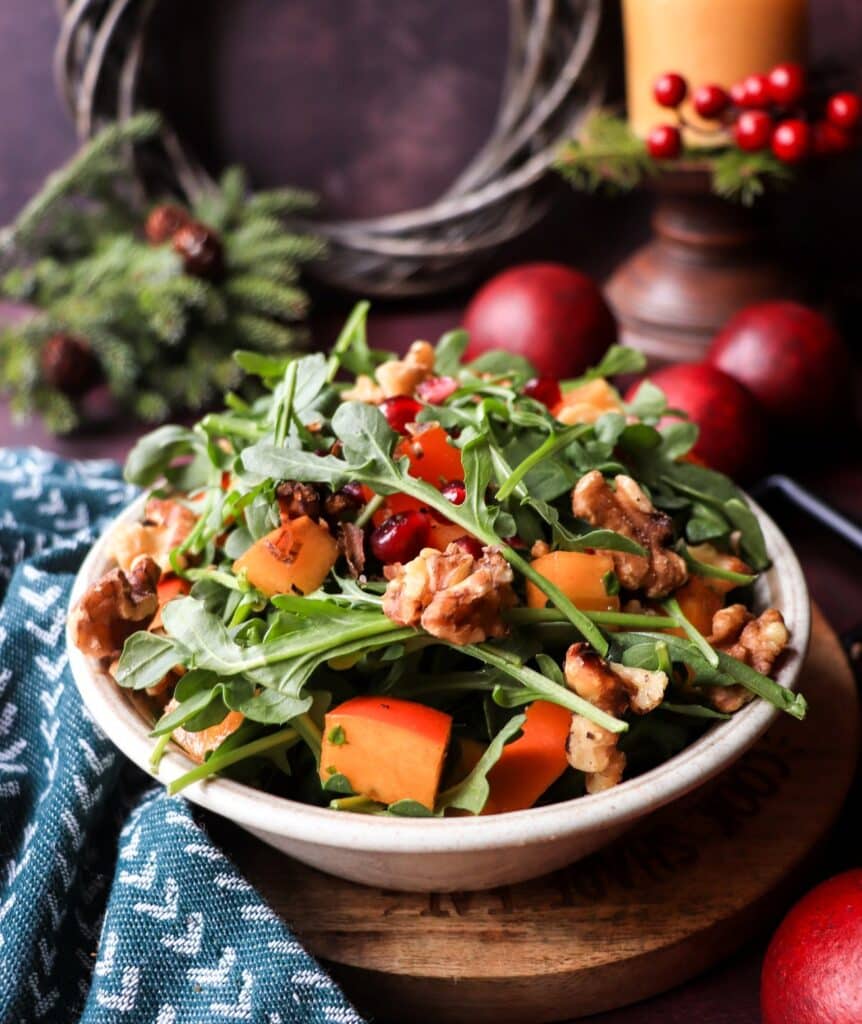 A festive Persimmon Pomegranate Walnut Salad with arugula, toasted walnuts, and bright orange persimmons, styled with holiday greenery and a candle in the background.