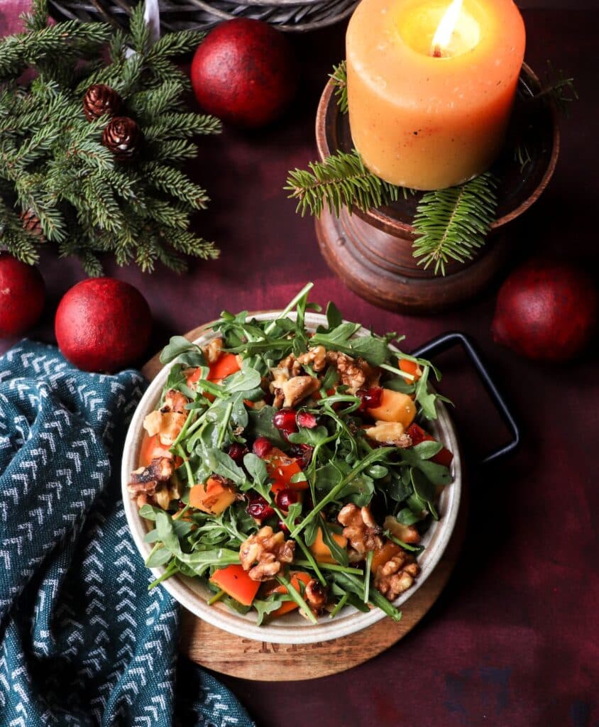 Overhead shot of Persimmon Pomegranate Walnut Salad with arugula, persimmons, walnuts, and pomegranate on a deep red background with pine branches and a burning candle — cozy holiday ambience.