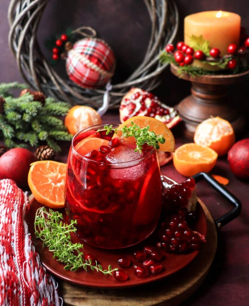 Festive pomegranate clementine holiday sangria served in a glass with pomegranate arils, clementine slices, and thyme, surrounded by holiday decor and warm candlelight.