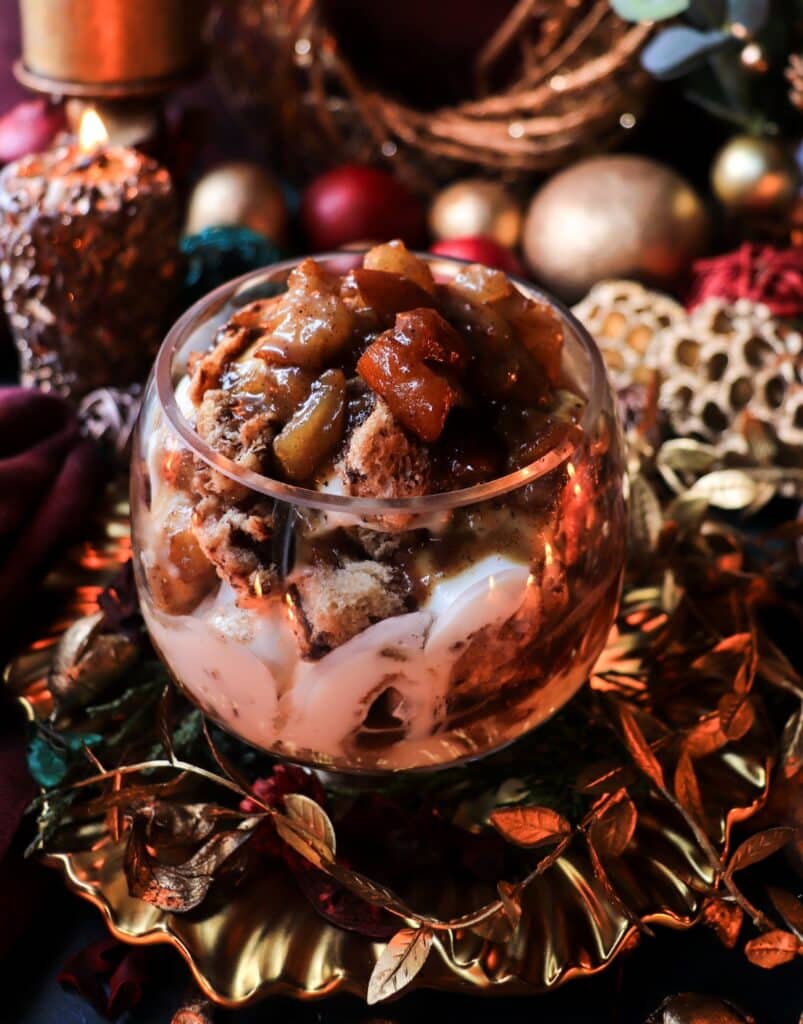 Festive caramelized brandy pear trifle layered with goat cheese cream and panettone, presented in a glass bowl with warm candlelight and holiday greenery in the background.