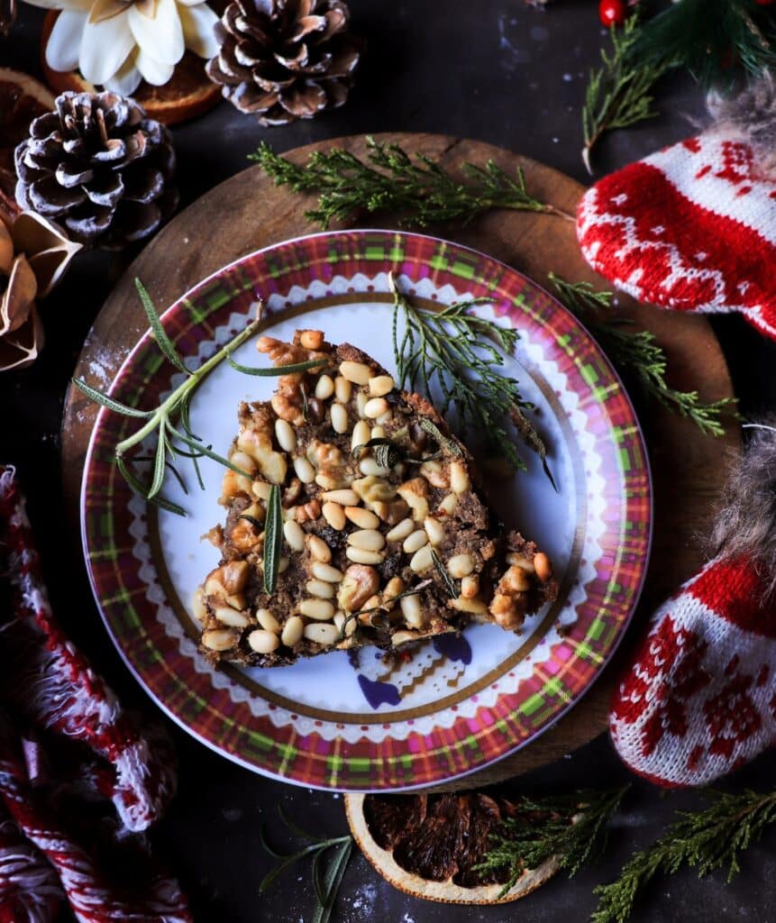 Slice of castagnaccio chestnut cake with pine nuts and walnuts served on a patterned plate, surrounded by pinecones, dried orange slices, evergreen sprigs, and cozy Christmas décor in a moody winter setting.