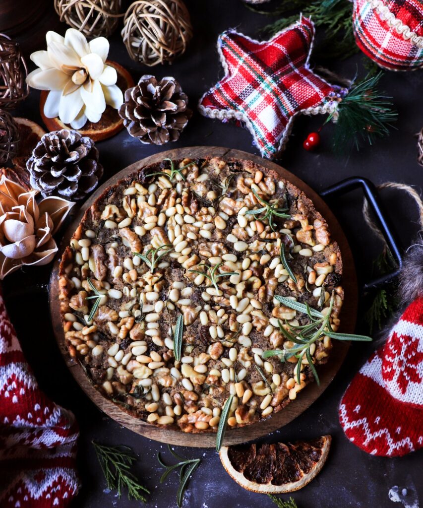 Whole Tuscan castagnaccio chestnut flour cake topped with pine nuts, walnuts, and rosemary, styled on a wooden board with pinecones, dried citrus slices, plaid fabric ornaments, and evergreen branches on a dark festive background.