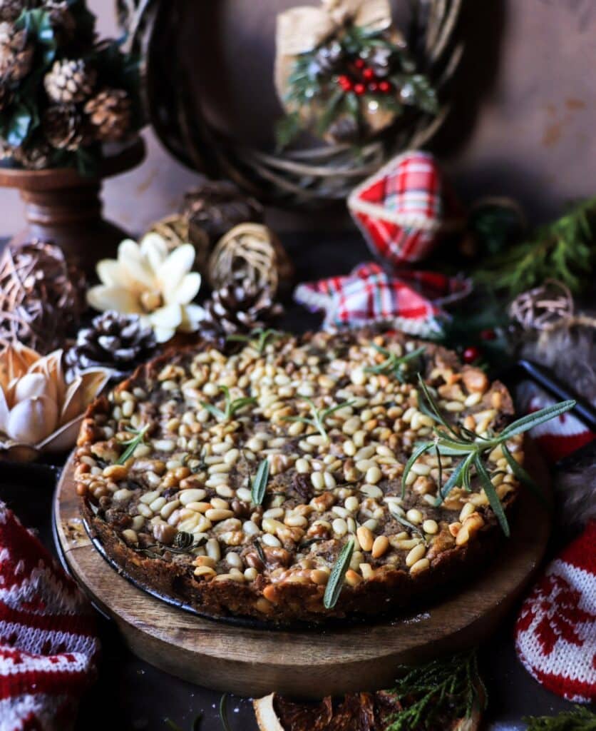 Rustic castagnaccio chestnut flour cake on a wooden stand, generously topped with pine nuts, walnuts, and rosemary, styled with pinecones, knitted red-and-white holiday textiles, and festive ornaments on a dark background.
