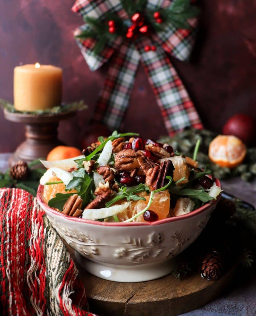 Overhead view of a bright clementine fennel salad with arugula, toasted pecans, and pomegranate arils, arranged in a red-rimmed bowl. Fresh clementines and a festive winter-knit cloth create a cheerful holiday atmosphere.
