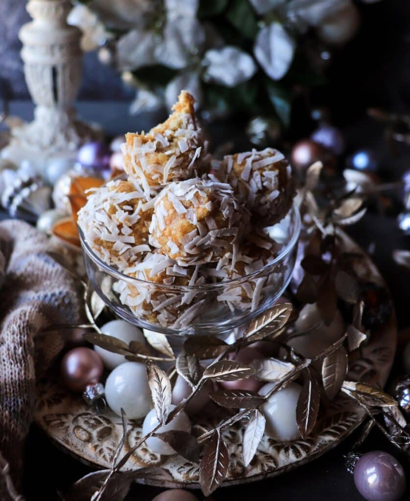 Close-up of coconut rum cookie truffles in a glass bowl, with one truffle showing a soft bite revealing the cashew–cookie filling, surrounded by festive ornaments and warm holiday décor