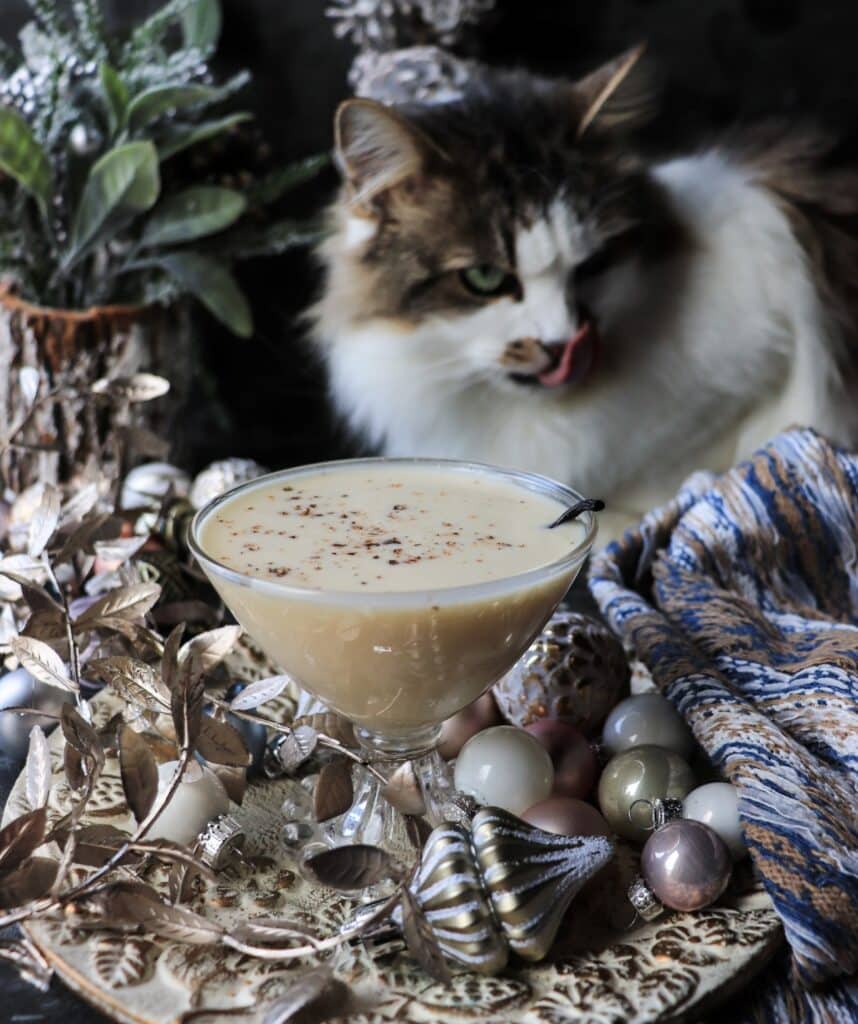 Creamy coquito holiday cocktail styled on a festive table with ornaments and winter greenery, with Daisy the cat curiously inspecting the drink in the background.