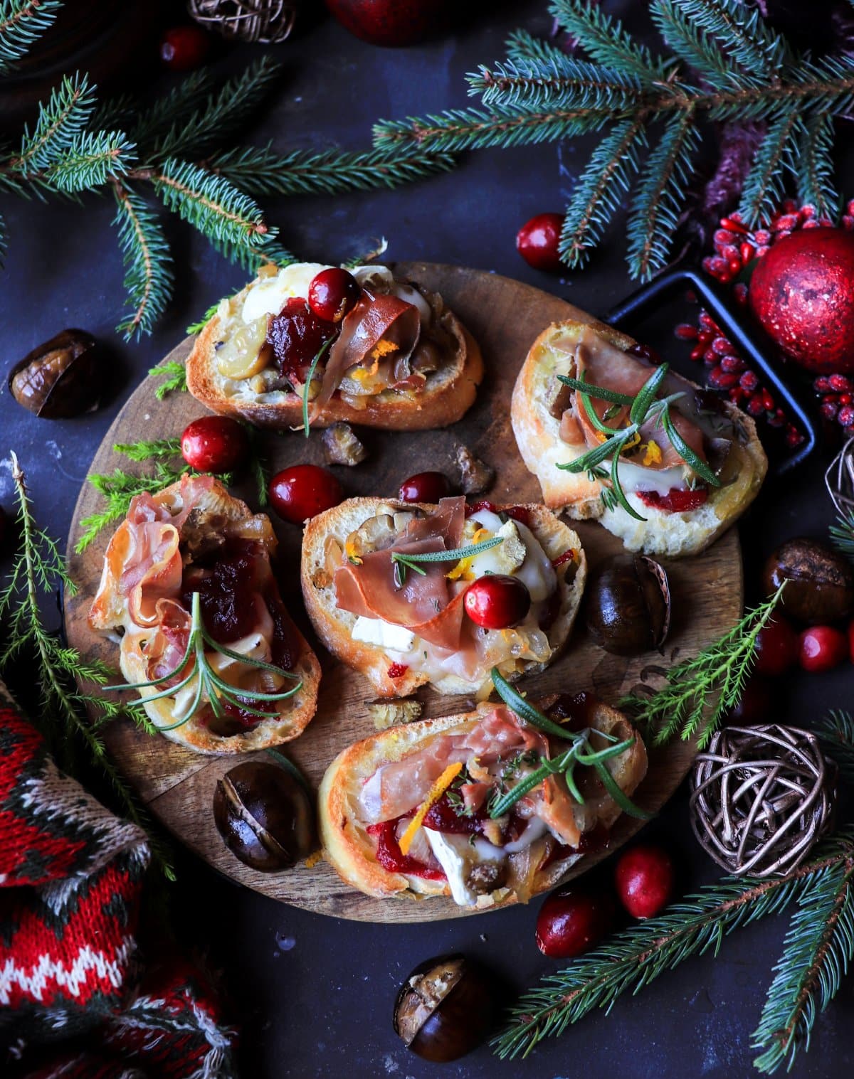 Festive cranberry chestnut brie crostini with prosciutto and caramelized onions, served on toasted baguette slices with rosemary, chestnuts, and warm candlelight in the background.