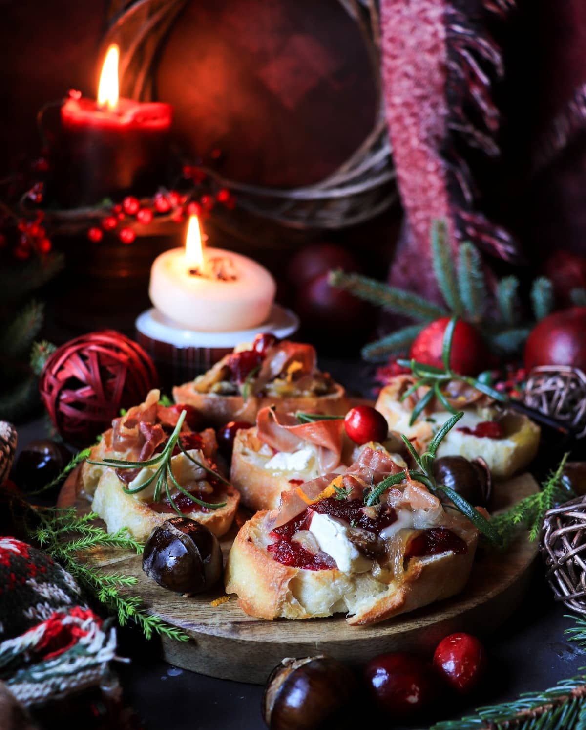 Cranberry chestnut brie crostini topped with prosciutto and caramelized onions, arranged on a wooden board with candles, evergreen branches, and festive red accents for a cozy holiday atmosphere.