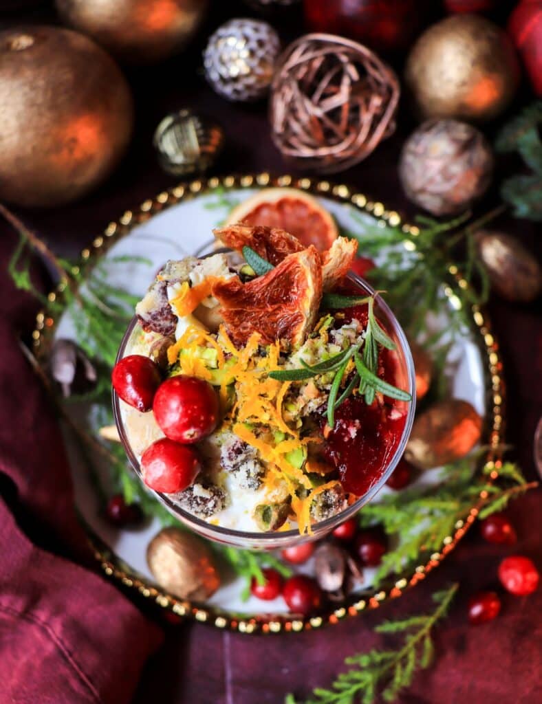 Close-up of a cranberry greek yogurt trifle showing vivid layers of yogurt, cranberry sauce, shortbread pieces, and pistachios, topped with orange zest, rosemary, and cranberries, surrounded by holiday greenery and metallic decorations