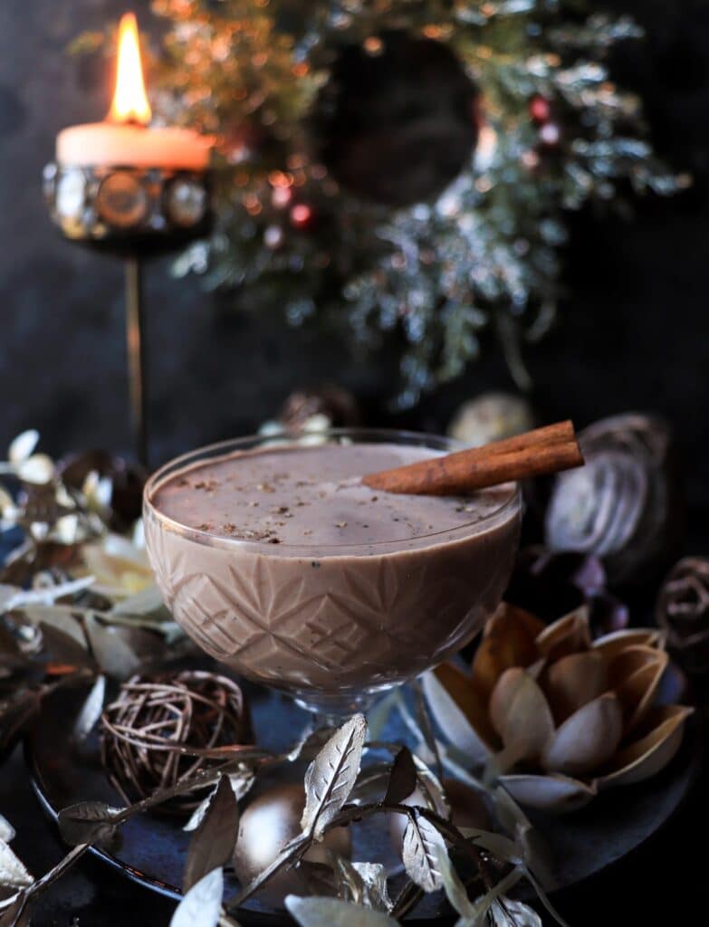 A festive eggnog brandy alexander cocktail served in a cut-glass coupe, topped with freshly grated nutmeg and garnished with a cinnamon stick, surrounded by metallic holiday décor. A glowing candle and blurred Christmas wreath create a warm, moody backdrop.