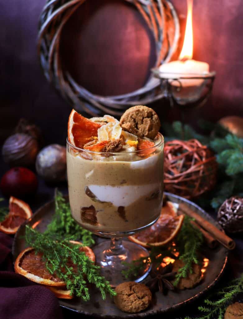 A layered gingerbread Greek yogurt trifle in a glass, showing the contrast of white and spiced yogurt with gingerbread cookie pieces and citrus garnish.
