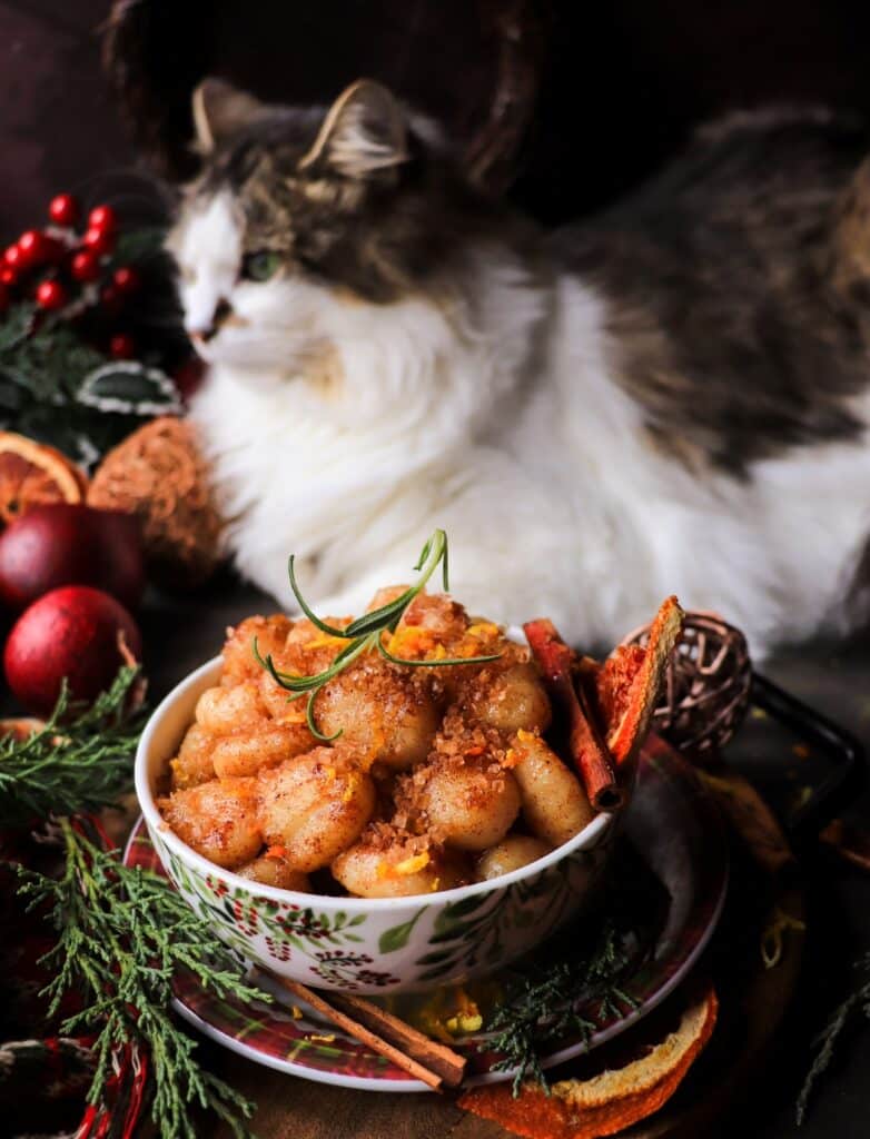 Fluffy white and tabby cat sitting behind a bowl of sweet skillet gnocchi topped with cinnamon, coarse sugar, and citrus zest, surrounded by festive Christmas greenery and spices.