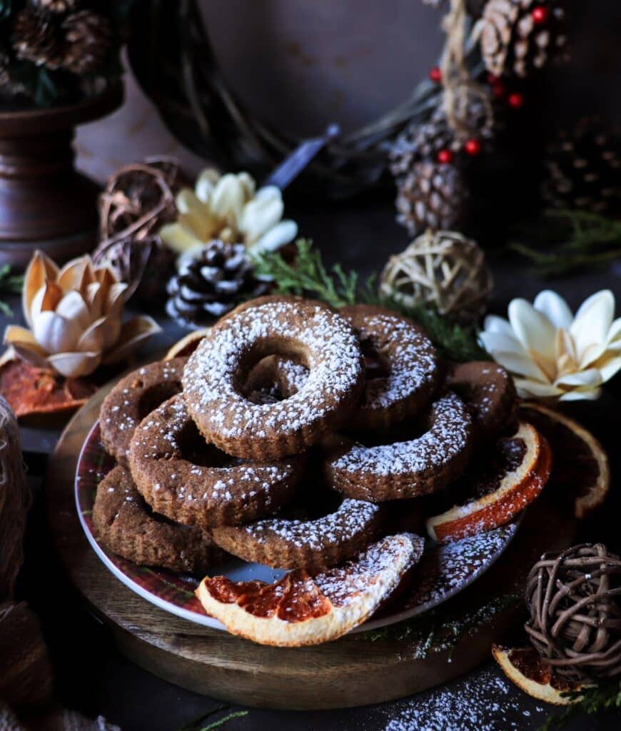 Gluten-free rye shortbread cookies dusted with powdered sugar, arranged on a festive plate with dried clementine slices, pinecones, and winter greenery