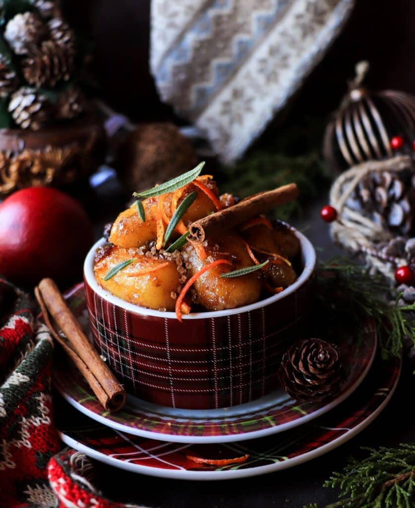 Soft dessert potato gnocchi coated in a glossy rum and raisin syrup, topped with cinnamon sticks, orange zest, and herbs, served in a red tartan ramekin on stacked festive plates, surrounded by pinecones, evergreen branches, yarn, and holiday décor.