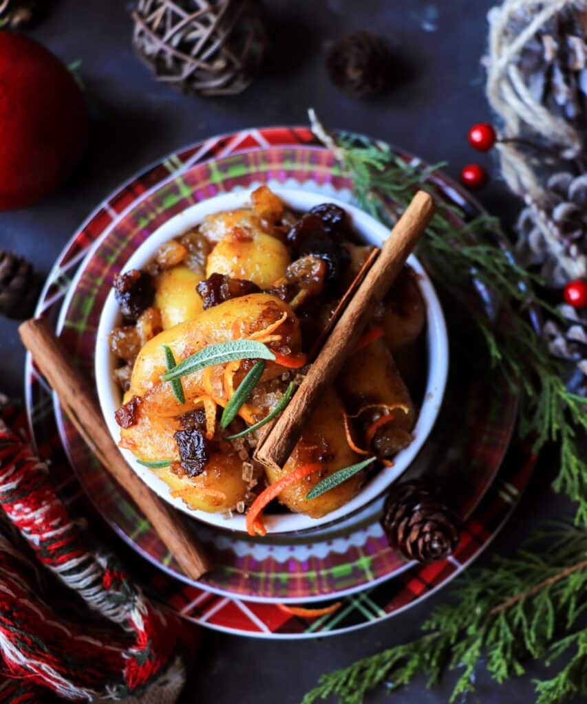 Overhead view of soft rum and raisin dessert gnocchi glazed in a sticky syrup, garnished with cinnamon sticks, citrus zest, and herbs, styled on plaid holiday plates with pinecones, evergreen sprigs, red berries, and textured winter décor.