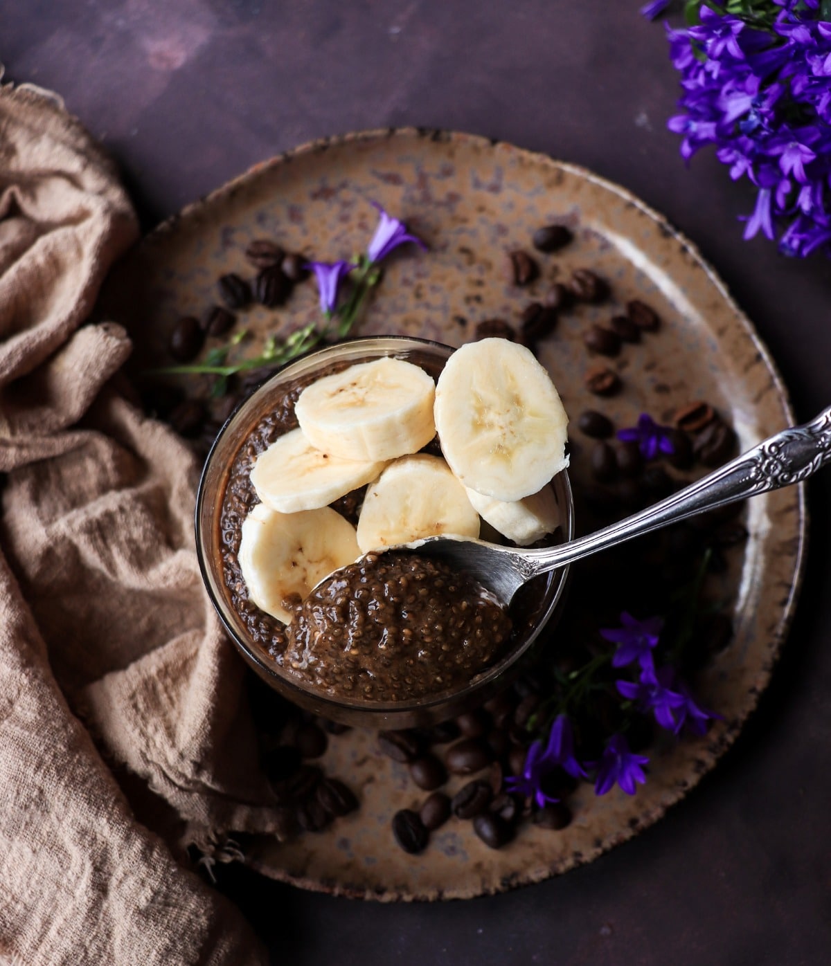 Banana coffee chia pudding in a glass, topped with fresh banana slices and shown with a spoon, coffee beans, and floral accents on a dark background.