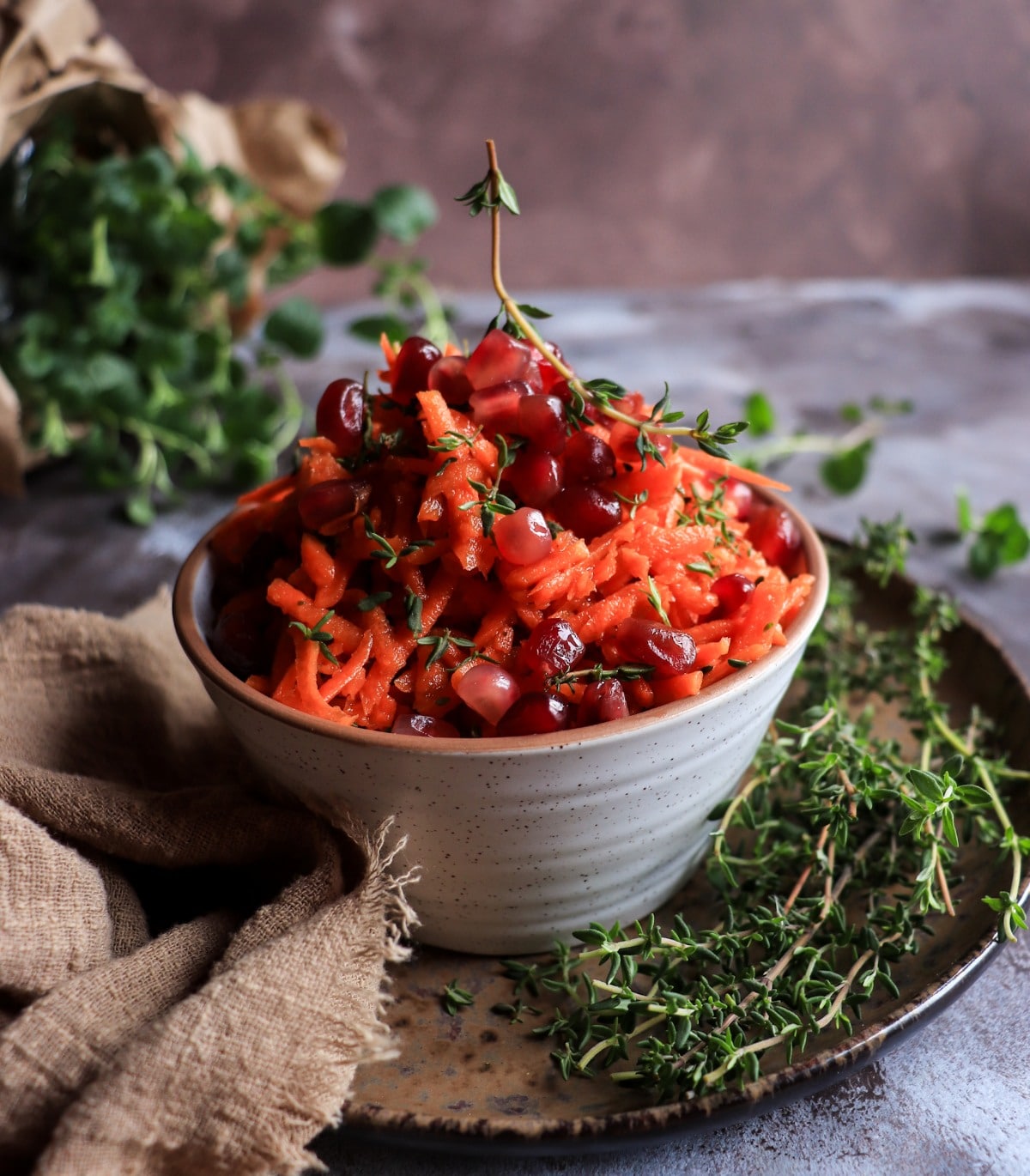 Shredded carrot salad with pomegranate molasses dressing, topped with juicy pomegranate arils and fresh thyme, served in a ceramic bowl.
