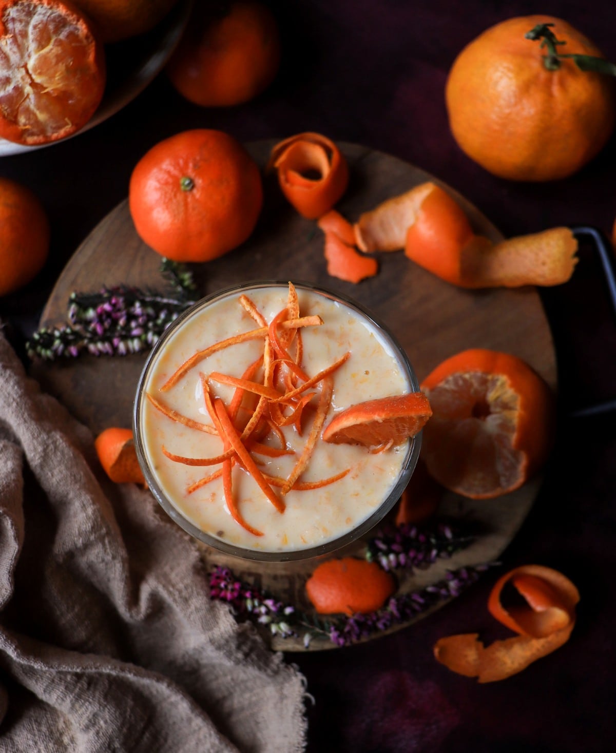 Overhead view of a clementine banana smoothie with Greek yogurt, garnished with clementine zest and a citrus slice, styled with whole and peeled clementines, citrus peels, and a dark textured background.