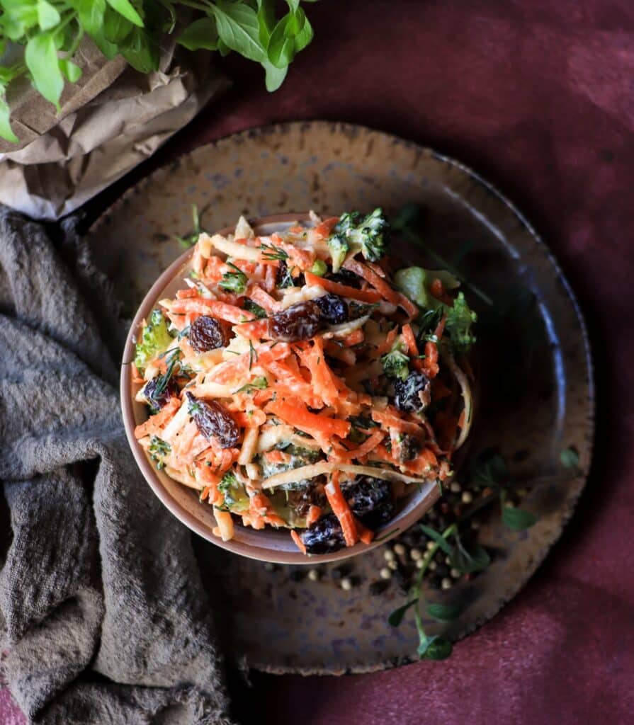 A close-up of raw broccoli, carrot, and parsnip salad with dried cranberries and raisins, mixed with a creamy yogurt-mayo dressing and fresh dill, served in a speckled bowl on a dark, moody background.