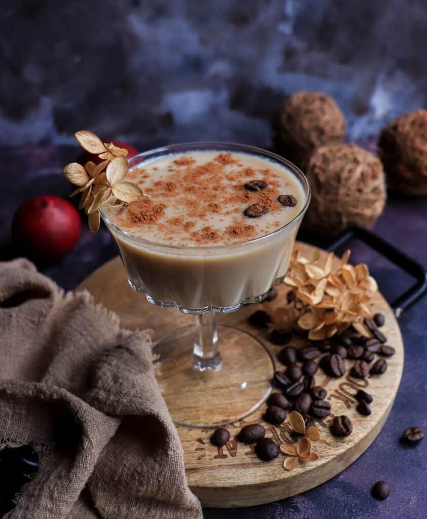 Creamy maple brandy coffee cocktail served in a coupe glass, dusted with cinnamon and garnished with coffee beans, styled with roasted coffee beans and dried flowers on a wooden board.
