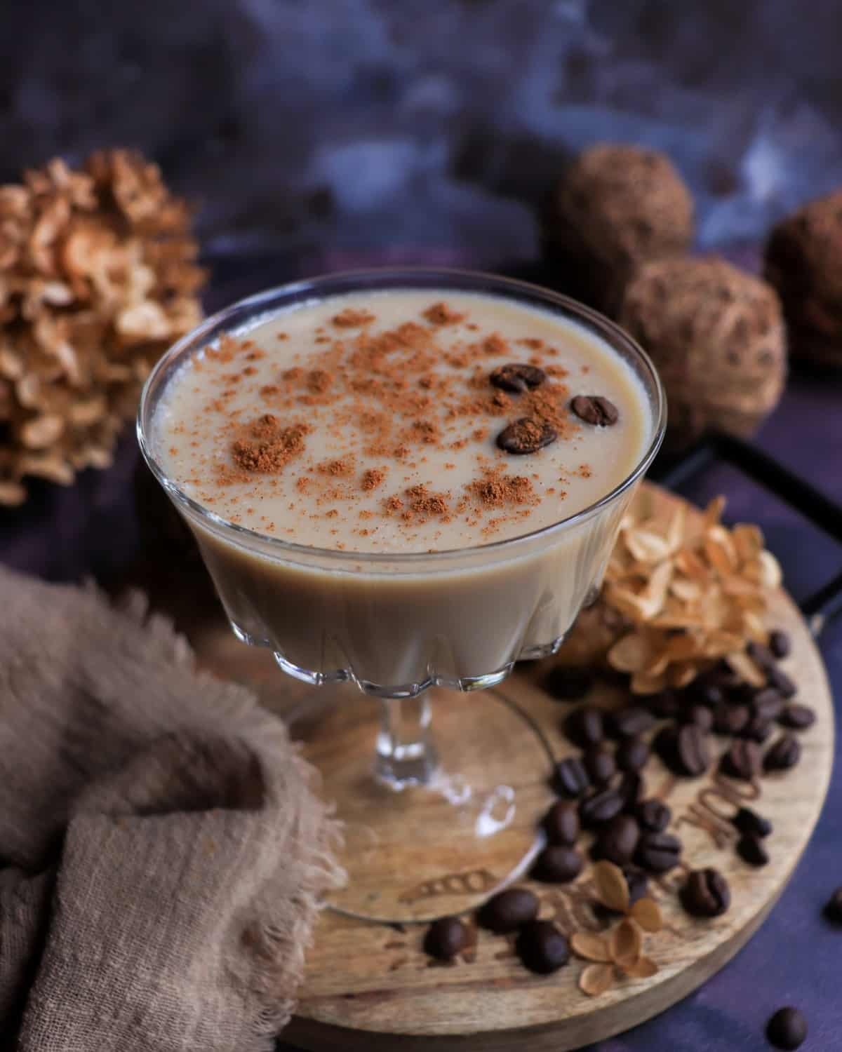 Creamy maple brandy coffee cocktail served in a coupe glass, dusted with cinnamon and garnished with coffee beans, styled with roasted coffee beans and dried flowers on a wooden board.