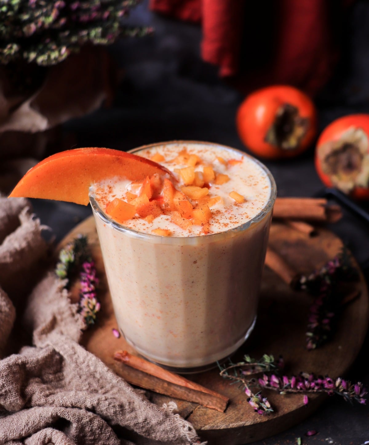 Creamy persimmon smoothie with Greek yogurt and cinnamon in a glass, topped with diced persimmon and a slice, with whole Fuyu persimmons in the background.