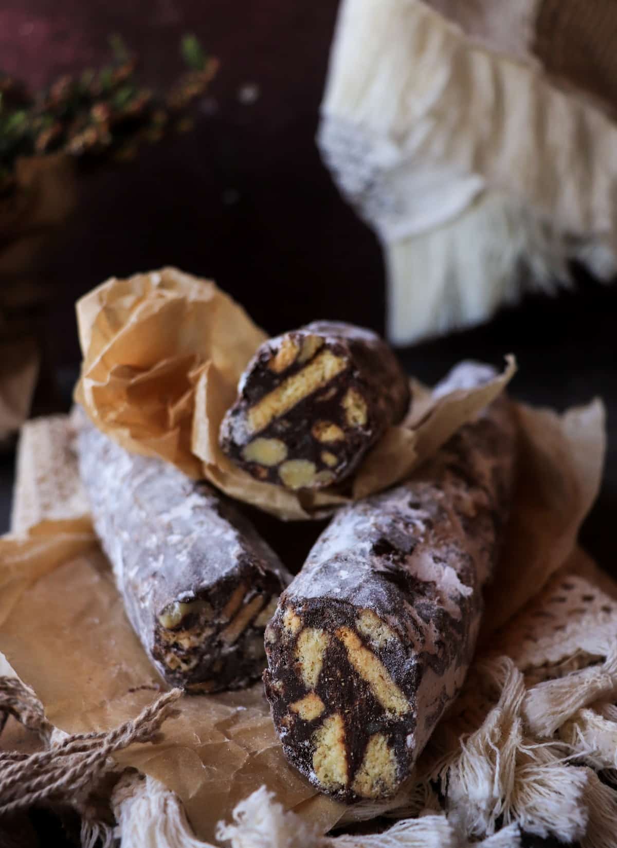 Italian chocolate salami logs dusted with powdered sugar, stacked on parchment with visible biscuit and nut mosaic inside