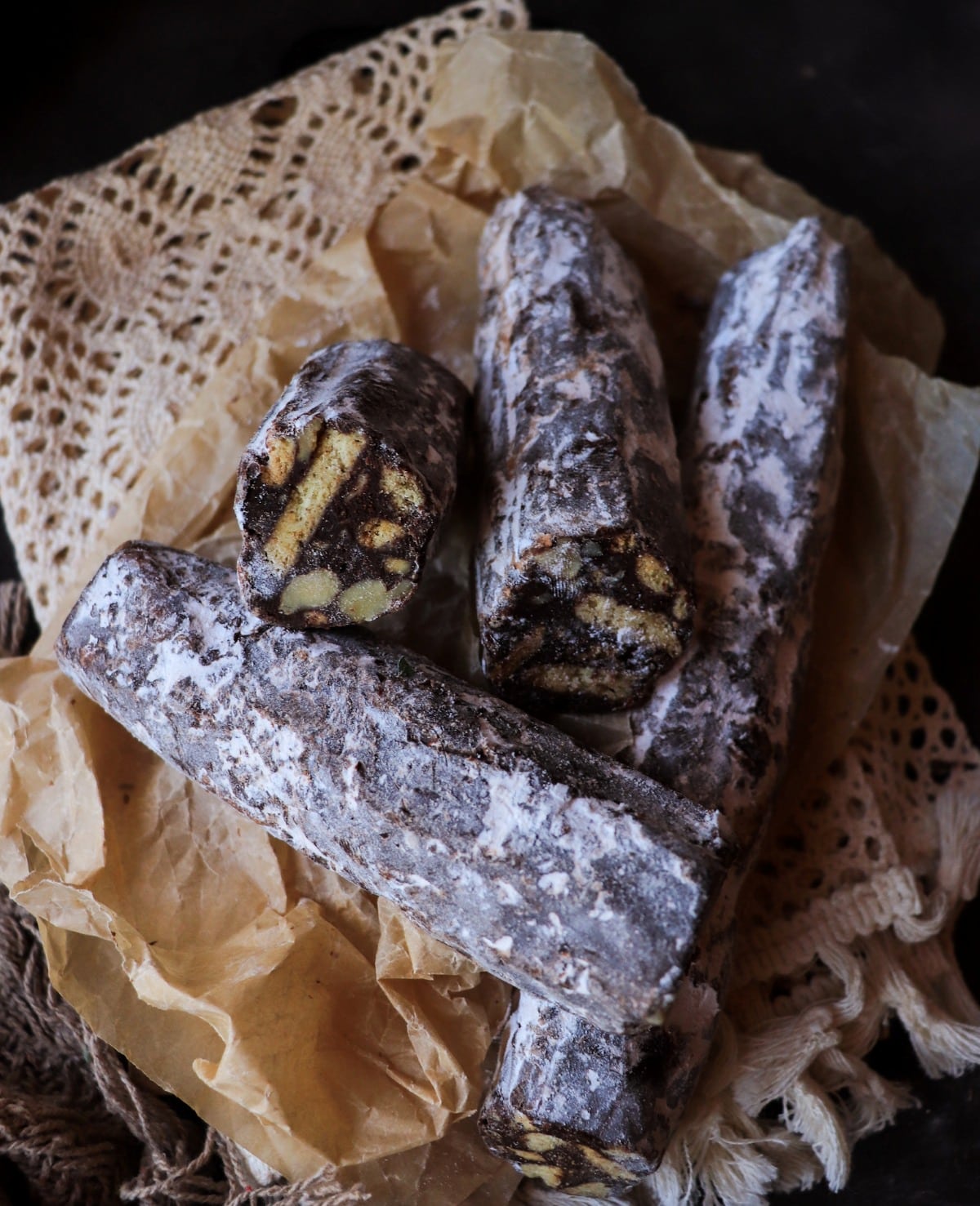 Italian chocolate salami logs dusted with powdered sugar, stacked on parchment with visible biscuit and nut mosaic inside